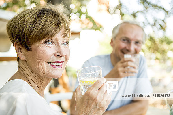 Lächelnde Seniorin trinkt ein Glas Wasser mit Ehemann im Hintergrund