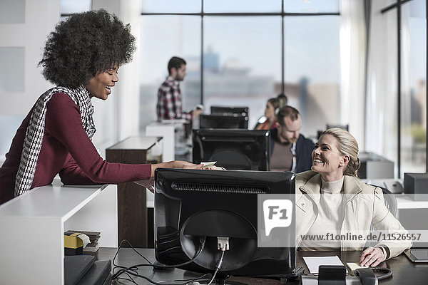 Beiläufige Büroangestellte übergibt Notizen an Mitarbeiterin hinter dem Schreibtisch mit Computer
