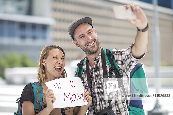 Young traveler couple taking pictures with sign saying 'Hi Mom'