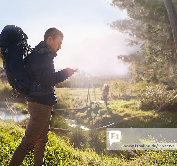 Young man with backpack hiking  checking smart phone in sunny field