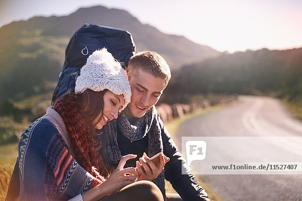 Young couple with backpack hiking  resting and using cell phone at sunny  remote roadside