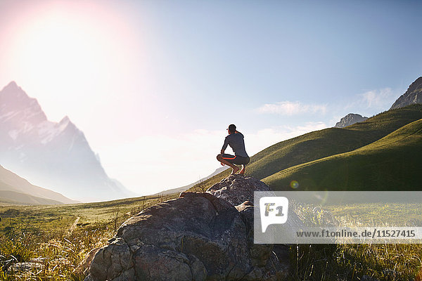 Young man crouching on rock  looking at sunny r emote mountain view