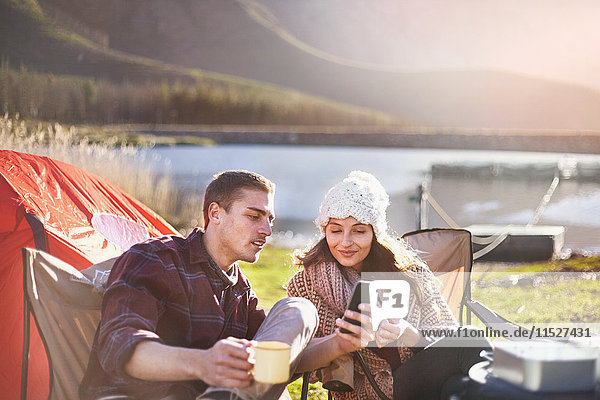 Young couple camping  drinking coffee and using cell phone at sunny lakeside campsite