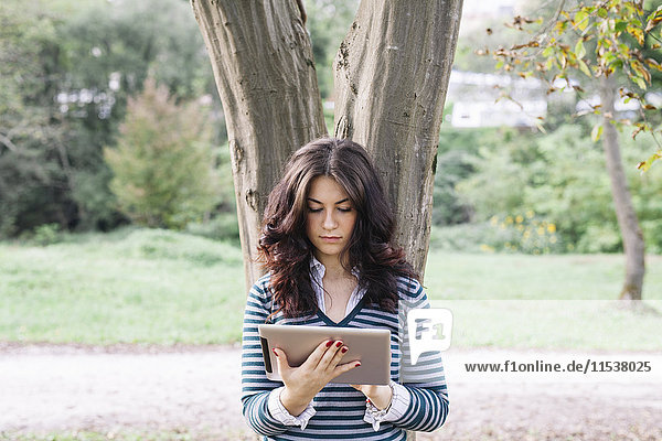 Junge Frau lehnt sich an den Baumstamm und schaut auf ein digitales Tablett.