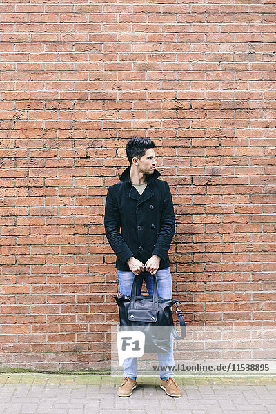 Young casual businessman leaning on brick wall carrying shoulder bag