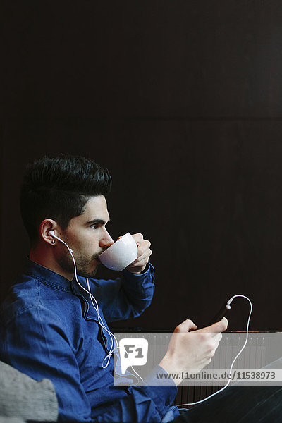 Profil eines jungen Mannes  der Musik mit Kopfhörern hört  während er vor schwarzem Hintergrund Kaffee trinkt.