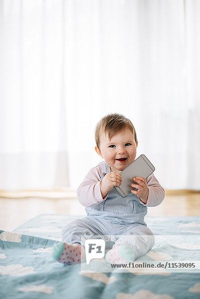 Portrait of smiling baby girl sitting on blanket holding smartphone