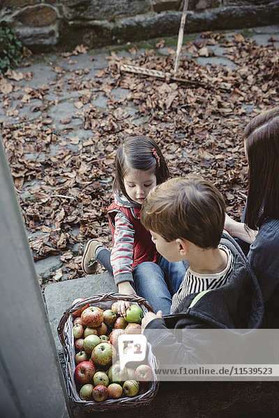 Frau sitzt am Eingang des Landhauses mit ihren Kindern und schaut auf einen Korb mit Äpfeln.