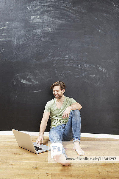 Porträt eines lächelnden jungen Mannes  der mit einem Laptop vor der Tafel auf dem Boden sitzt.