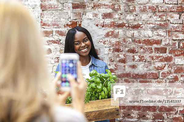 Smiling young woman holding crate with herbs being photographed