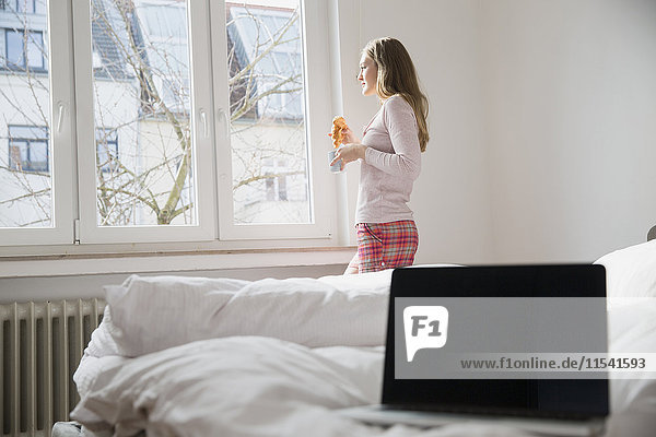 Junge Frau mit Croissant und Tasse Kaffee stehend im Schlafzimmer mit Blick durchs Fenster