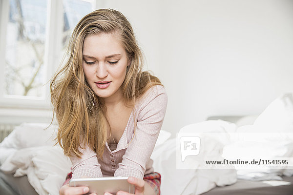 Junge Frau sitzt auf ihrem Bett und schaut auf das Smartphone.
