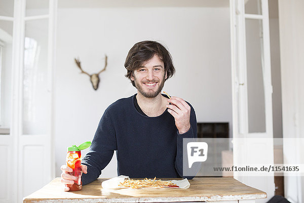 Porträt eines lächelnden jungen Mannes beim Essen von Pommes frites mit Ketchup