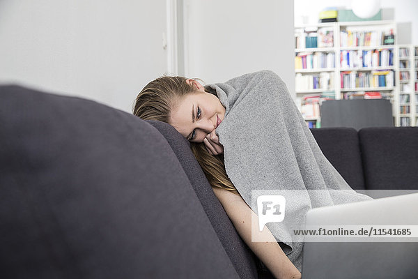 Daydreaming young woman covered with blanket sitting on the couch at home
