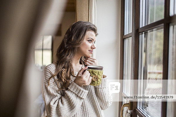 Junge Frau zu Hause mit Blick aus dem Fenster bei einer Tasse Kaffee