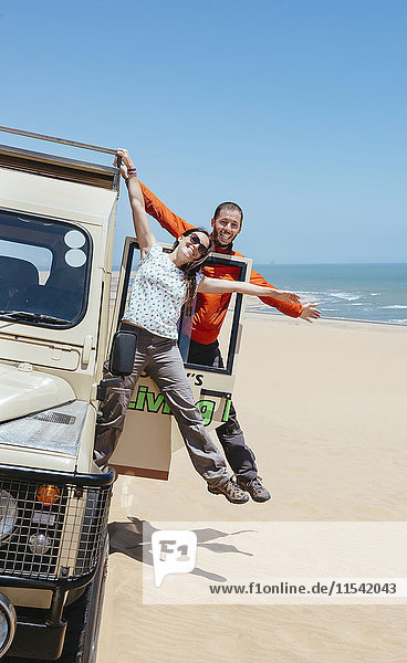 Namibia  Namib Wüste  Swakopmund  glückliches Paar  das in der Wüste an einem Geländewagen hängt  mit dem Meer im Hintergrund