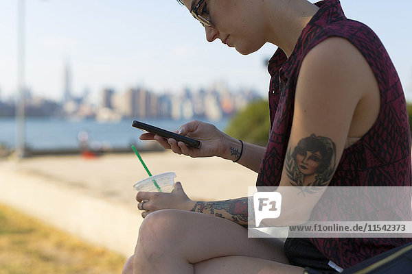 USA  New York City  Brooklyn  tätowierte Frau sitzt auf einer Bank und schaut auf ihr Smartphone.