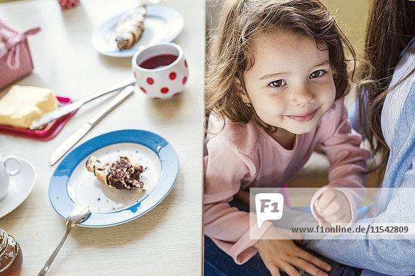 Porträt eines lächelnden kleinen Mädchens  das mit seiner Mutter am Frühstückstisch kuschelt.