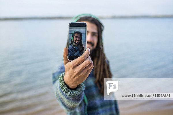 Junger Mann mit Dreadlocks nimmt Selfie mit einem Smartphone am Strand.