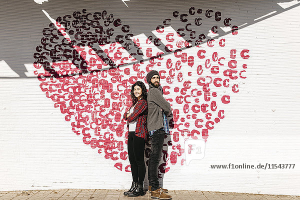 Junges Paar steht Rücken an Rücken vor einer Backsteinmauer mit Herz