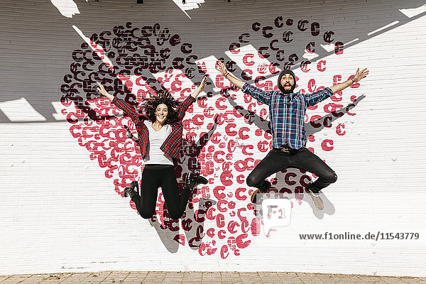 Ein glückliches junges Paar springt vor eine Ziegelmauer mit Herz