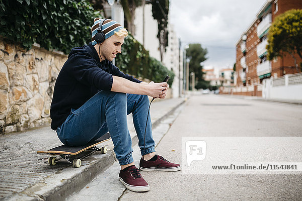 Spain  Torredembarra  smiling young man sitting on his skateboard at curb listening music with headphones