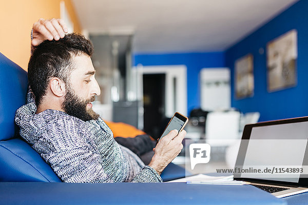 Bearded young man working at home relaxed sitting on the couch  using laptop and mobile