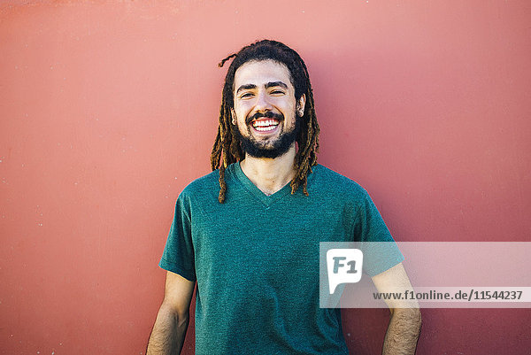 Porträt eines lachenden jungen Mannes mit Dreadlocks und Bart vor einer roten Wand