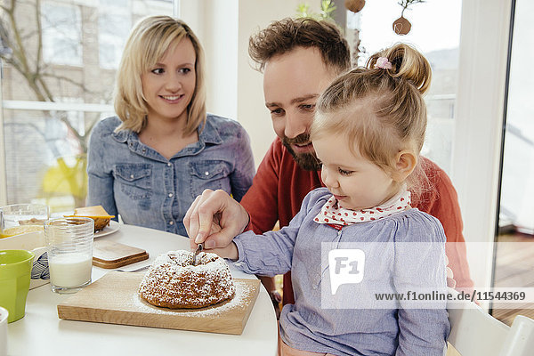 Kleines Mädchen schneidet Kuchen mit Hilfe ihres Vaters  während die Mutter sie beobachtet.
