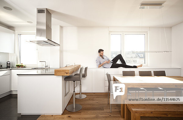Mann auf Sideboard liegend in seiner offenen Küche mit Smartphone