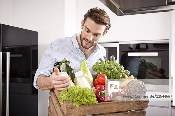 Portrait of young man with wooden box of fresh vegetables and two milk bottles in his kitchen