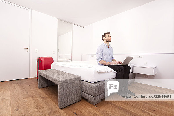 Mann sitzend mit Laptop auf einem Hotelbett