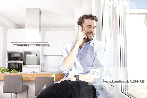 Junger Mann sitzt auf der Fensterbank in seiner Küche und schaut durchs Fenster  während er telefoniert.