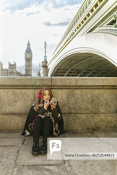 UK  London  young woman listening music and painting her lips near Westminster Bridge