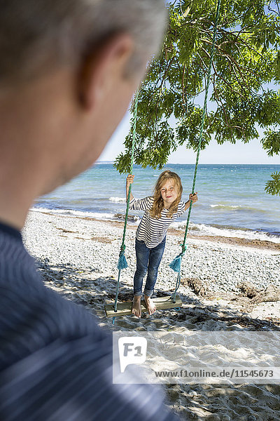 Lächelndes Mädchen steht auf einer Schaukel am Strand und schaut ihren Vater im Vordergrund an.