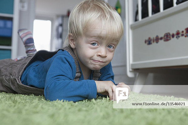 Porträt eines lustigen kleinen blonden Jungen  der zu Hause auf einem grünen Teppich liegt.