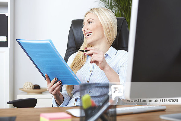 Lächelnde blonde Frau sitzt am Schreibtisch und hält Dokumente.