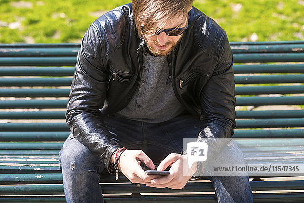Mann mit Sonnenbrille  der mit seinem Smartphone auf der Parkbank sitzt.