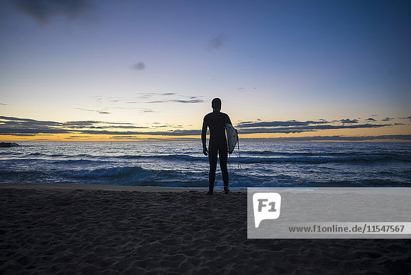 Rückansicht des am Strand stehenden Surfers bei Sonnenaufgang