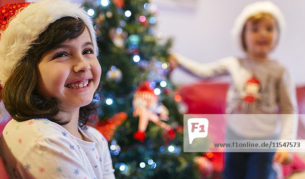 Porträt eines glücklichen Mädchens zur Weihnachtszeit mit ihrer kleinen Schwester im Hintergrund