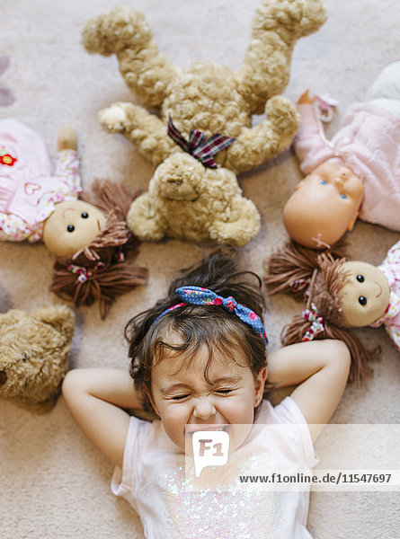 Porträt eines lachenden kleinen Mädchens auf dem Boden liegend mit Teddys und Puppen um sie herum.