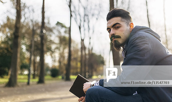 Porträt des bärtigen jungen Mannes mit Buch im Park