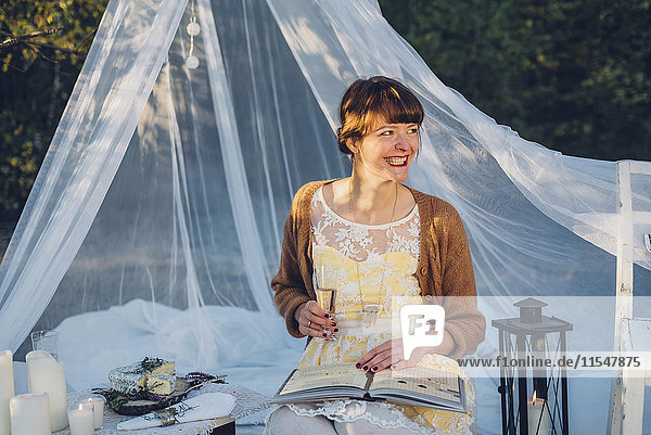Porträt einer lachenden Frau mit Buch und einem Glas Champagner in einem romantischen Camp in der Natur.