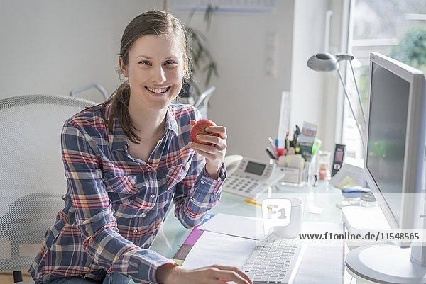 Porträt einer lächelnden jungen Frau am Schreibtisch im Büro  die einen Apfel hält.