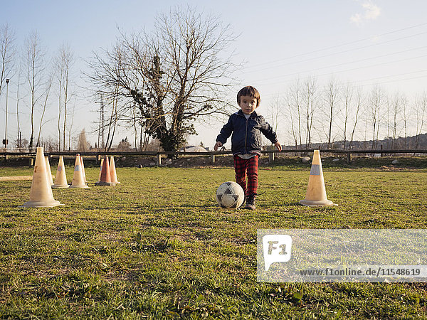 Kleiner Junge spielt Fußball auf Rasen zwischen Verkehrskegeln