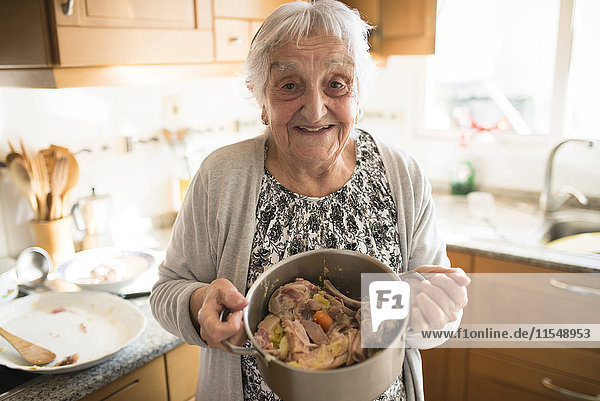 Portrait of smiling senior woman showing a cooking pot of Galician stew in the kitchen
