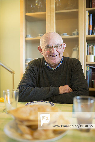 Porträt eines lächelnden älteren Mannes  der zu Hause am gedeckten Tisch sitzt.