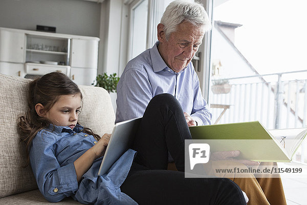 Großvater und seine Enkelin sitzen zusammen auf der Couch mit Buch und digitalem Tablett.