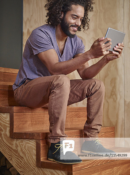Smiling young man sitting on wooden stairs looking at digital tablet