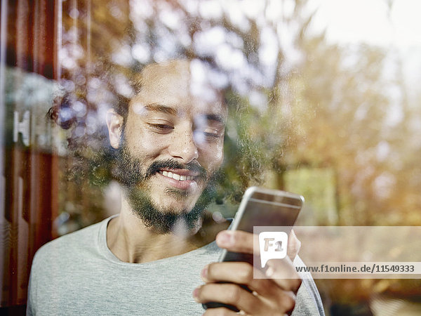 Smiling young man looking on cell phone behind windowpane